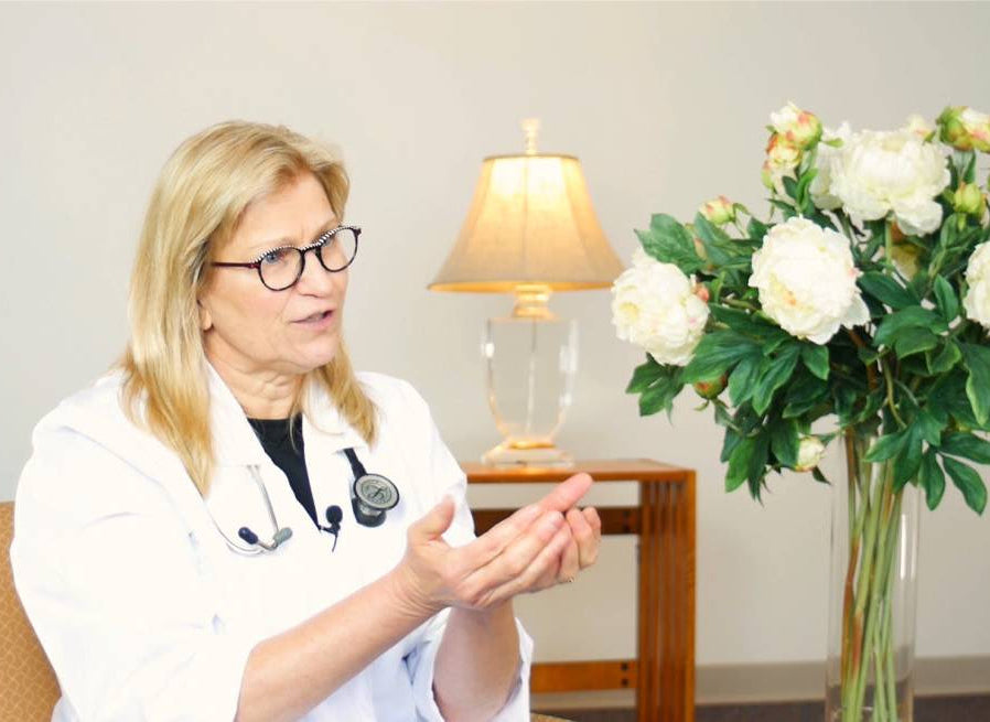 Doctor in a white coat sitting in a chair with a vase of flowers on a table behind her.